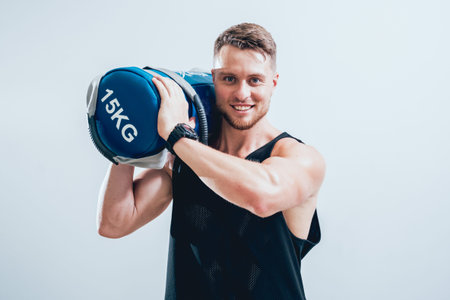Athletic young man training with sandbags at gray background. Crossfit center