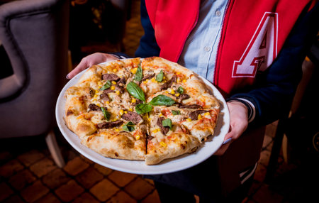 Waiter, carrying dish with tasty pizza with chicken and vegetables. Restaurant. Background