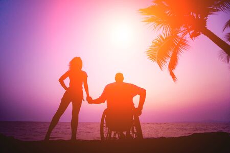 Disabled man in a wheelchair with his wife on the beach. Silhouettes at sunset. Conceptの写真素材