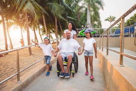 Disabled man in a wheelchair moves on a ramp to the beach with his family.の写真素材