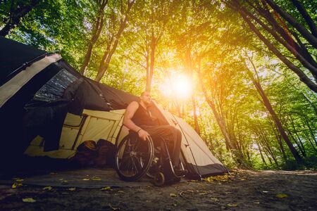 Disabled man resting in a campsite with friends. Wheelchair in the forest on the background of tents. Camping. Summertimeの写真素材