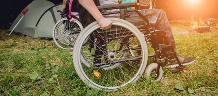 Disabled man resting in a campsite with friends. Wheelchair in the forest on the background of tents. Camping. Summertimeの写真素材
