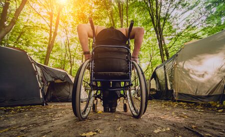 Disabled man resting in a campsite with friends. Wheelchair in the forest on the background of tents. Camping. Summertimeの写真素材