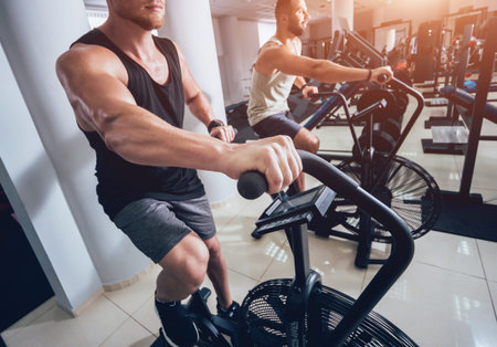 Young men with muscular body using air bike for cardio workout at cross training gym. Fitness...