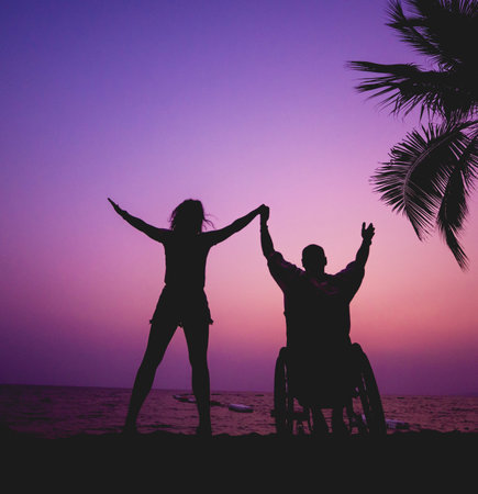Disabled man in a wheelchair with his wife on the beach. Silhouettes at sunset