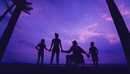 Disabled man in a wheelchair with his family on the beach. Silhouettes at sunsetの写真素材