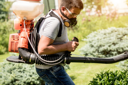 Gardener in protective mask and glasses spraying toxic pesticides trees and bushes. Landscape...