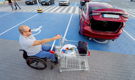 Adult disabled man in a wheelchair puts purchases in the trunk of a car in a supermarket parking lotの写真素材
