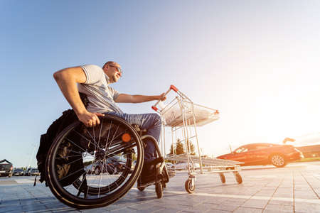 Adult disabled man in a wheelchair pushes a cart towards a car in a supermarket parking lotの写真素材