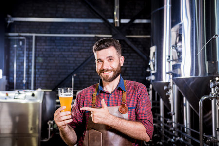 Young brewer wearing a leather apron is testing beer at a modern brewery