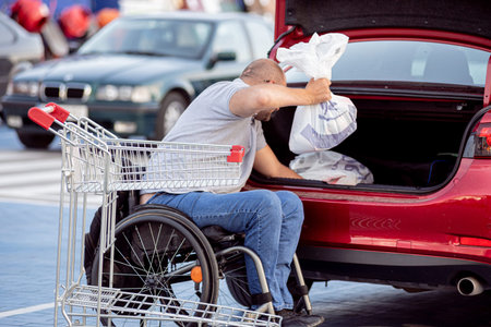 Adult disabled man in a wheelchair puts purchases in the trunk of a car in a supermarket parking lot