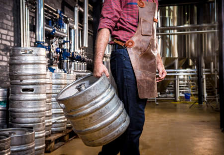 Young male brewer in leather apron holds barrel with craft beer at modern brewery factoryの写真素材