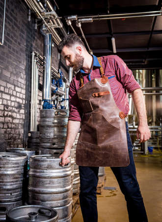 Young male brewer in leather apron holds barrel with craft beer at modern brewery factoryの写真素材