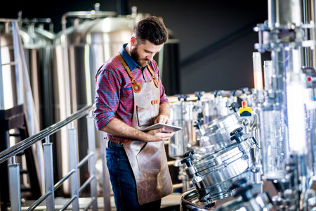 Young male brewer in leather apron supervising the process of beer fermentation at modern brewery...