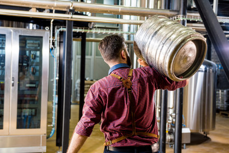 Young male brewer in leather apron holds barrel with craft beer at modern brewery factory