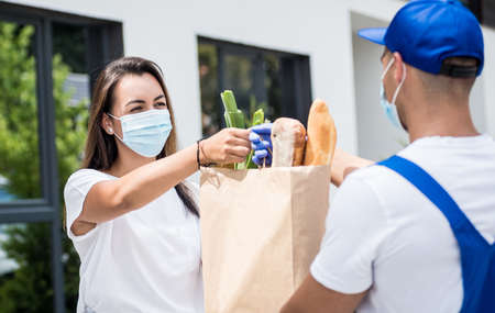 Young courier wearing a protective mask and gloves delivers goods to a young woman during quarantineの写真素材