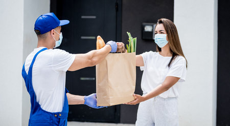 Young courier wearing a protective mask and gloves delivers goods to a young woman during quarantineの写真素材