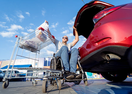 Adult disabled man in a wheelchair puts purchases in the trunk of a car in a supermarket parking lotの写真素材