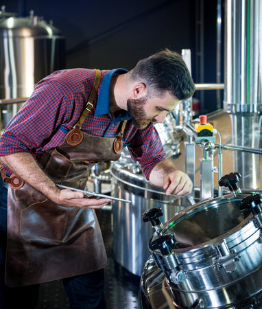 Young male brewer in leather apron supervising the process of beer fermentation at modern brewery...