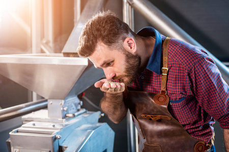 A young brewer in a leather apron controls the grinding of malt seeds in a mill at a modern brewery