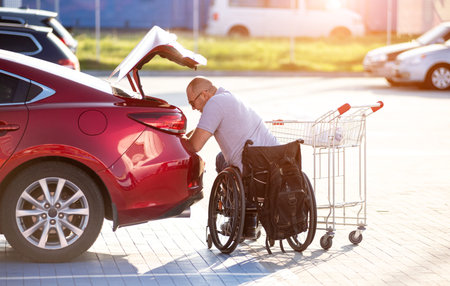 Adult disabled man in a wheelchair puts purchases in the trunk of a car in a supermarket parking lotの写真素材
