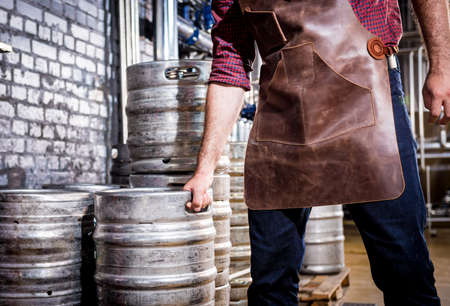 Young male brewer in leather apron holds barrel with craft beer at modern brewery factoryの写真素材