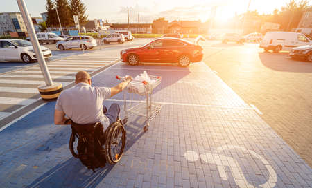 Adult disabled man in a wheelchair pushes a cart towards a car in a supermarket parking lotの写真素材