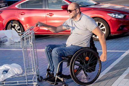 Disabled man in wheelchair pushing cart in front of himself at supermarket parkingの写真素材