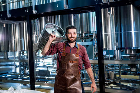 Young male brewer in leather apron holds barrel with craft beer at modern brewery factoryの写真素材