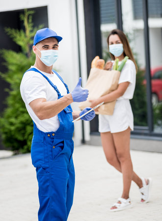 Young courier wearing a protective mask and gloves delivers goods to a young woman during quarantine