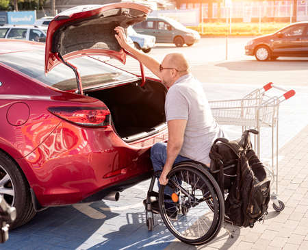 Adult disabled man in a wheelchair puts purchases in the trunk of a car in a supermarket parking lotの写真素材