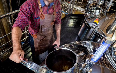 Young male brewer in leather apron supervising the process of beer fermentation at modern brewery...