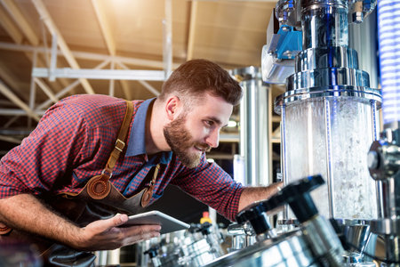 Young male brewer in leather apron supervising the process of beer fermentation at modern brewery...
