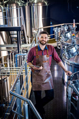 Young male brewer in leather apron supervising the process of beer fermentation at modern brewery factoryの写真素材