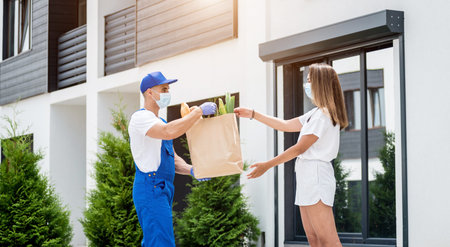 Young courier wearing a protective mask and gloves delivers goods to a young woman during quarantine
