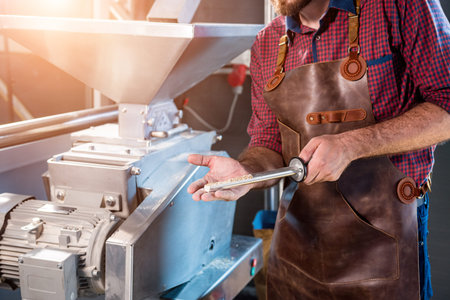 A young brewer in a leather apron controls the grinding of malt seeds in a mill at a modern brewery