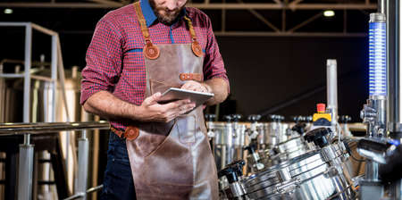 Young male brewer in leather apron supervising the process of beer fermentation at modern brewery factoryの写真素材