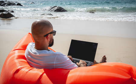 Young man working on his laptop on the beach while sitting on an inflatable sofaの写真素材