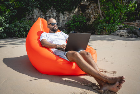 Young man working on his laptop on the beach while sitting on an inflatable sofa