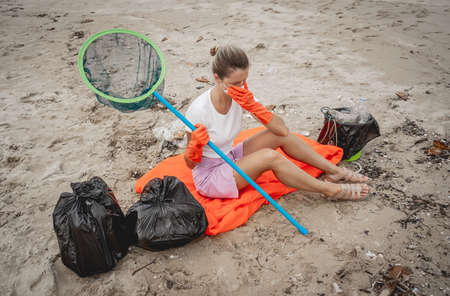 A female ecologist volunteer is resting after cleaning the beach on the seashore from plastic and other wasteの写真素材