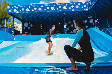 Young man surfing with trainer on a wave simulator at a water amusement park