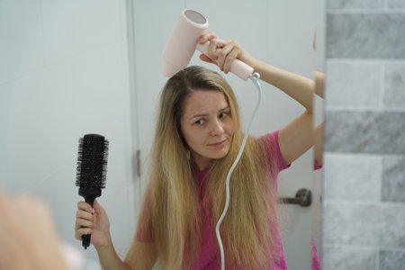 A woman is expertly styling her hair using a blow dryer and brush while in the bathroomの写真素材