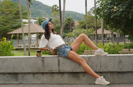 A young woman sits comfortably on a low wall at the park holding a skateboard in her hands