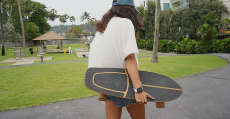 A woman is walking at the city park while carrying a skateboard in her hands