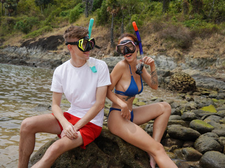 A young couple wear goggles and snorkels siting on a rock by the waterの写真素材