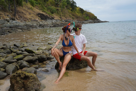 A young couple wear goggles and snorkels siting on a rock by the waterの写真素材