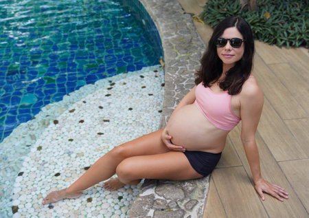 A pregnant woman in a pink bikini sits by a swimming pool