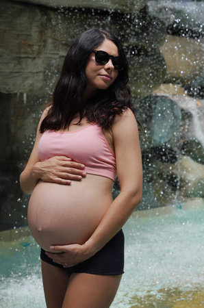 A pregnant woman is gracefully standing in front of a beautiful waterfall rock