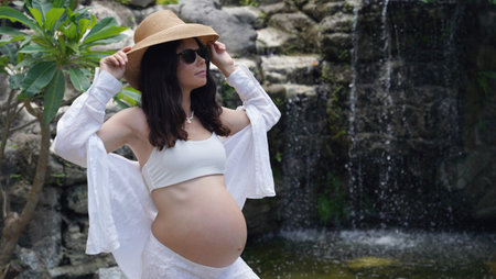 A pregnant woman is gracefully standing in front of a beautiful waterfall rockの写真素材
