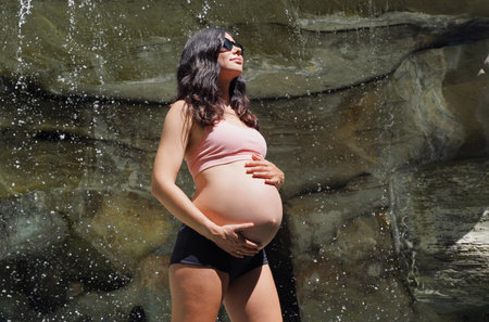 A pregnant woman is gracefully standing in front of a beautiful waterfall rockの写真素材